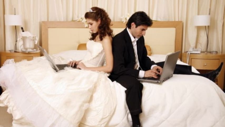 moglie e marito seduti sul letto di spalle mentre scrivono al computer 