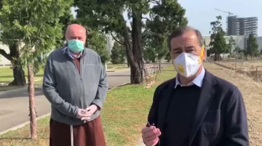 Beppe Sala con la mascherina e il microfono in mano di fronte al cimitero di Milano. Alla sua destra, leggermente indietro, anche un parroco