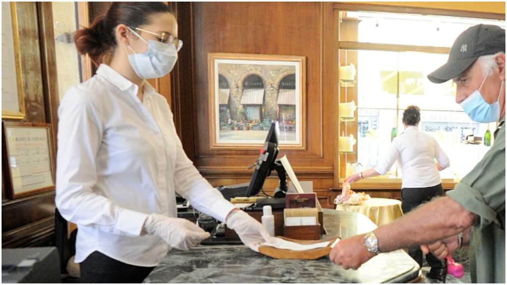 una barista alla cassa con mascherina e guanti che passa una ricevuta a un cliente, con la mascherina e il cappellino