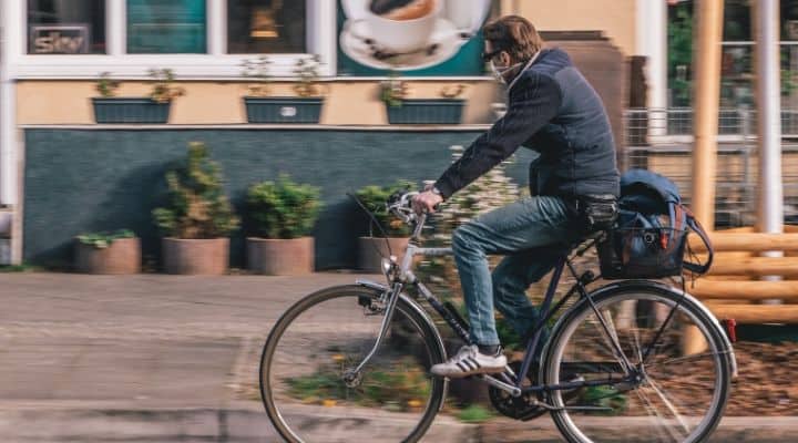 Un uomo che va in bicicletta. Sullo sfondo una casa e alcuni vasi di fiori davanti alla facciata