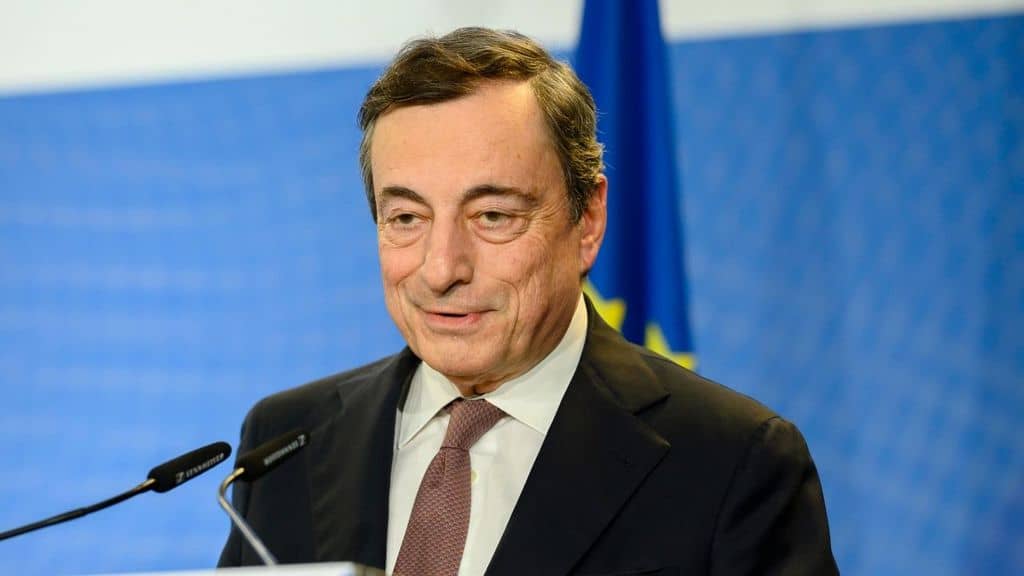 Mario Draghi di fronte a un microfono, su sfondo bianco e bliu